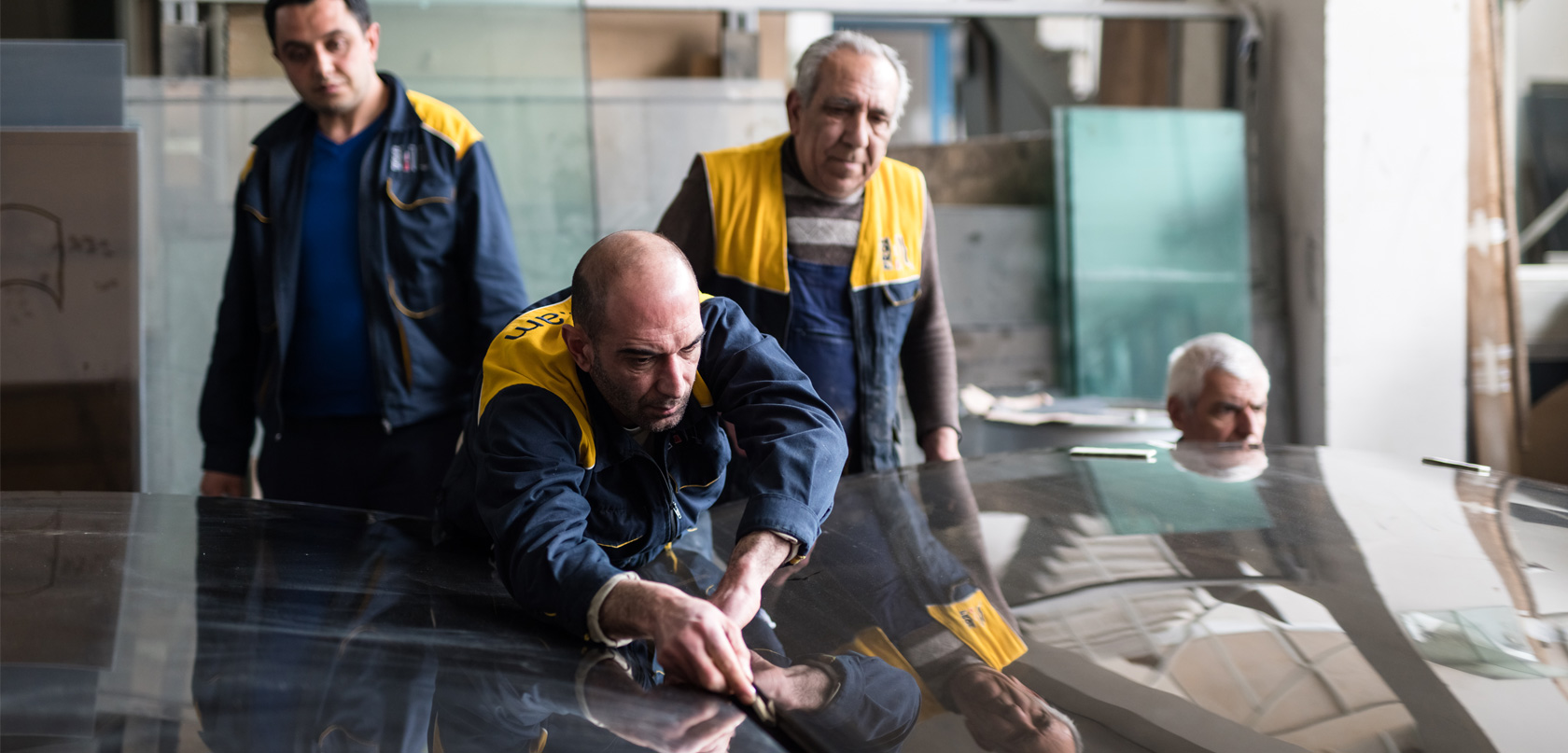 Trois hommes en veste d'uniforme jaune et bleu marine travaillent en usine. L’un d’eux se penche sur une feuille de plastique posée sur une table. 